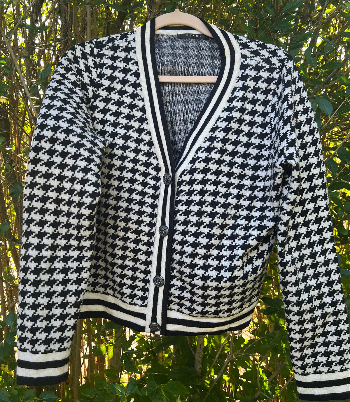 A black and white houndstooth cardigan sweater hanging on a wooden hanger against a background of green foliage.