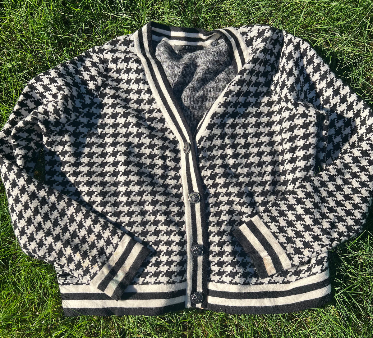 A black and white houndstooth cardigan jacket with striped ribbed trim, laid flat on green grass.