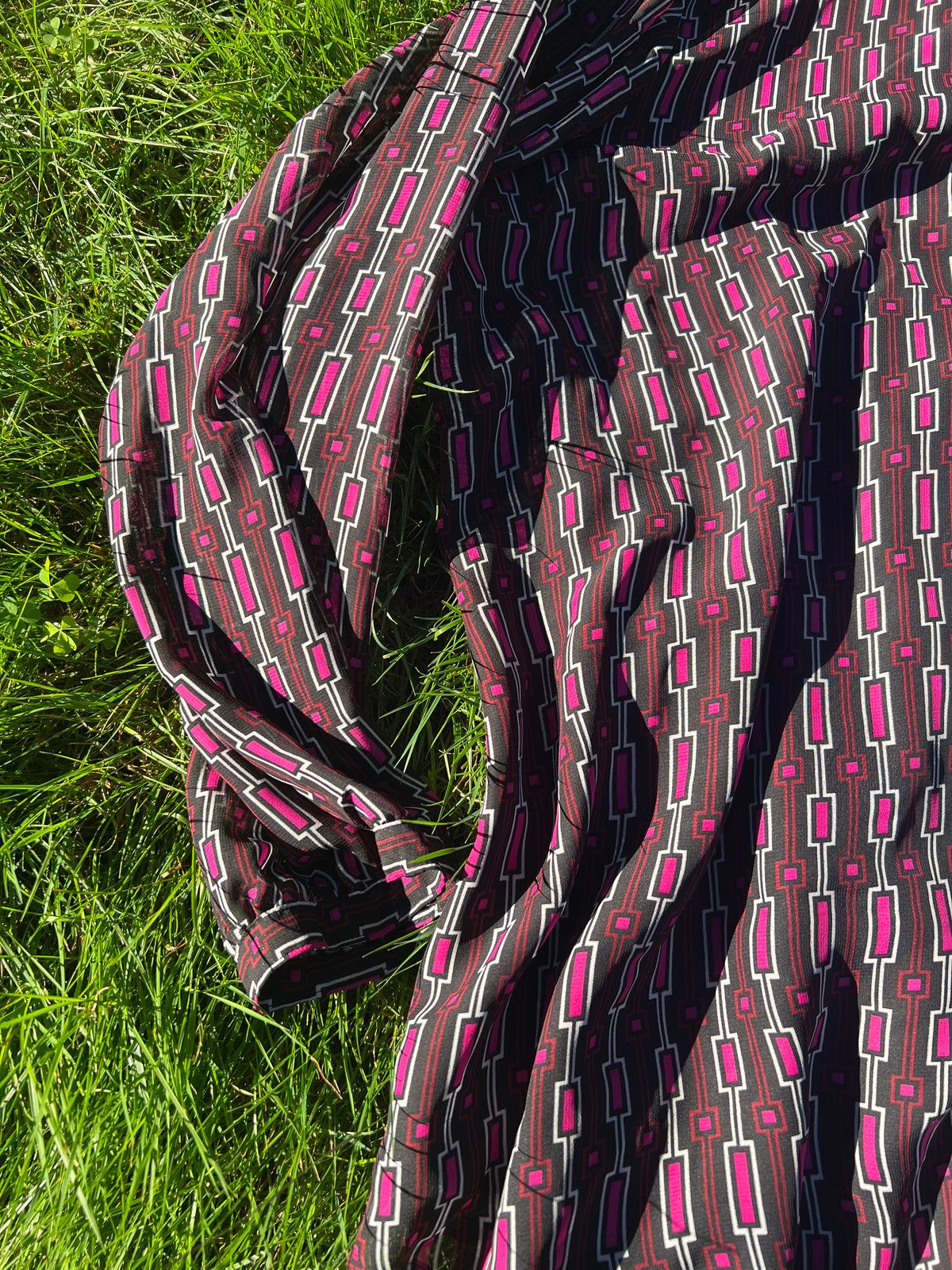 A draped piece of patterned fabric with a brown, white, and bright pink geometric design, laid out on green grass.