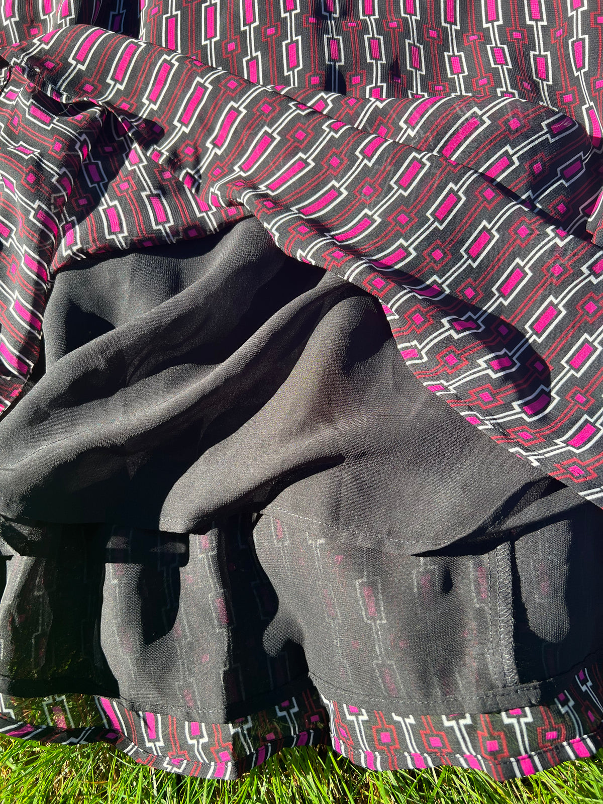 A close-up of a draped fabric garment or textile with a dark charcoal gray front panel and a geometric patterned section in black, white, and bright pink, laid outdoors on grass in direct sunlight.