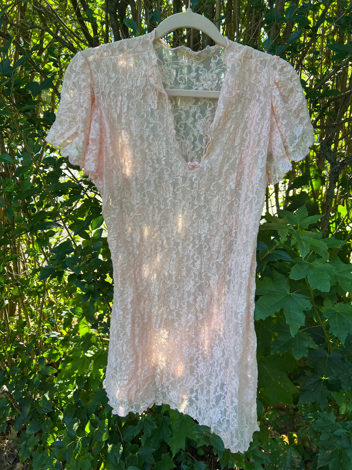 A vintage pale blush pink lace short-sleeve top hanging on a white plastic hanger against a backdrop of green outdoor foliage.