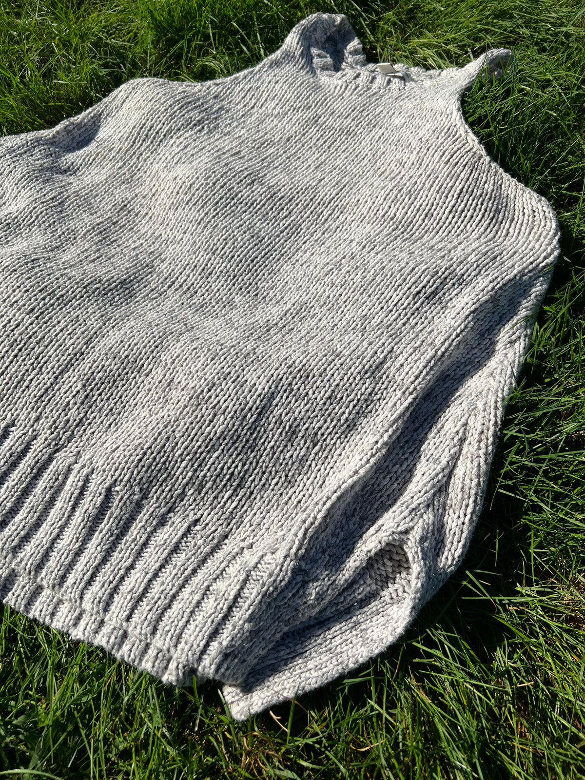 A light gray hand-knit sleeveless top with a ribbed hem, laid flat on green grass.