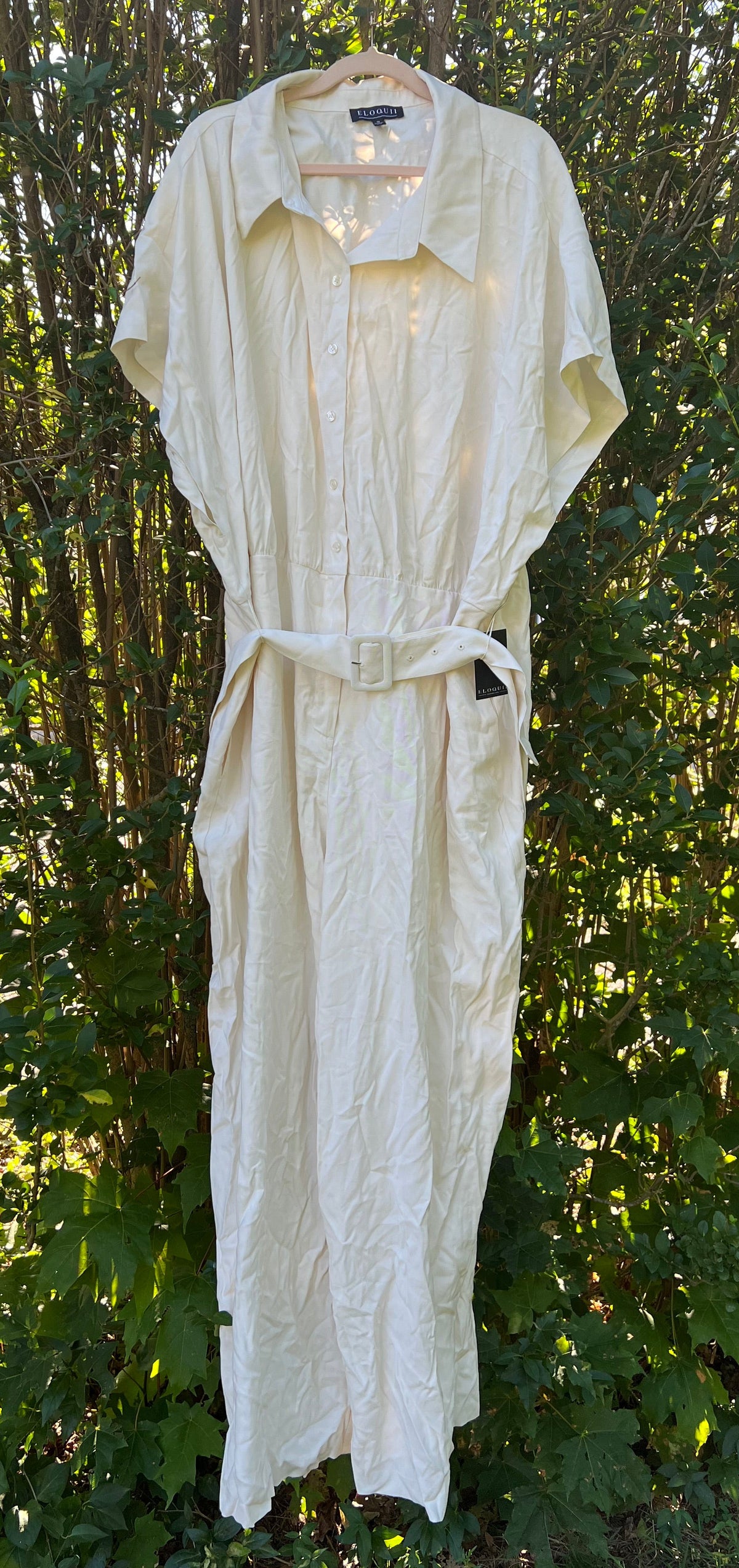 A cream-colored linen shirt Jumpsuit with a matching tie belt, hanging on a hanger against a backdrop of green bushes and trees.